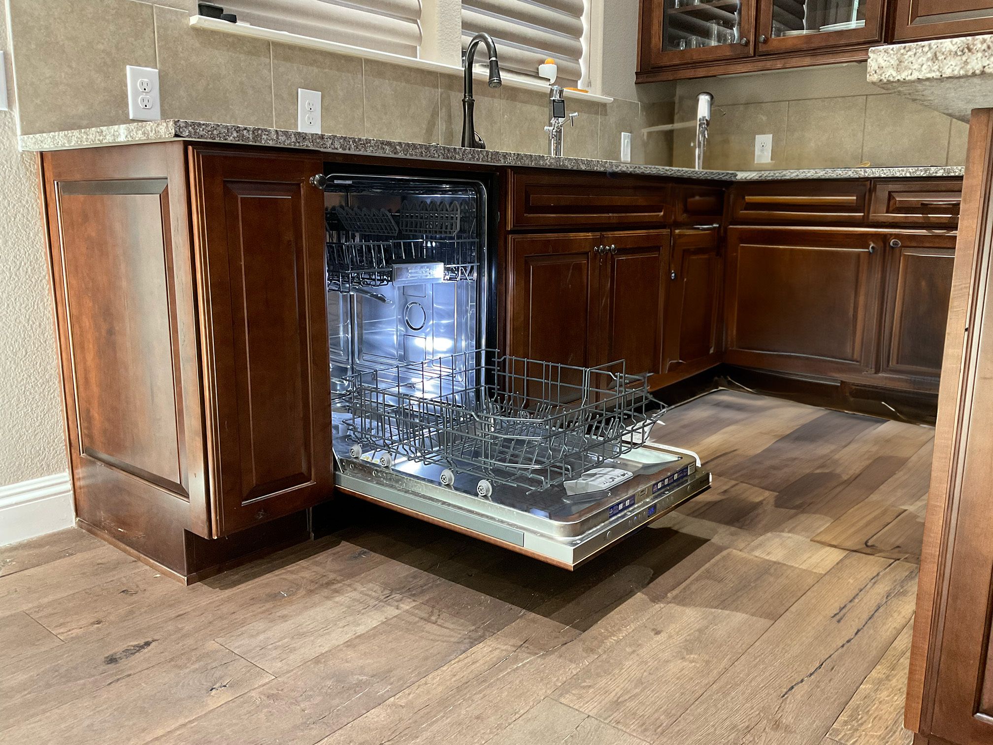 A kitchen scene featuring an open dishwasher with a silver interior and rack, set against wooden cabinets and a granite countertop.