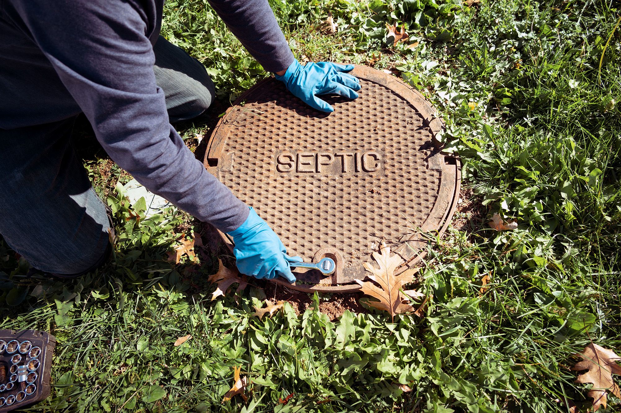 Inspector in blue gloves kneels by a grassy area, using a wrench to open a septic tank manhole cover labeled "SEPTIC."
