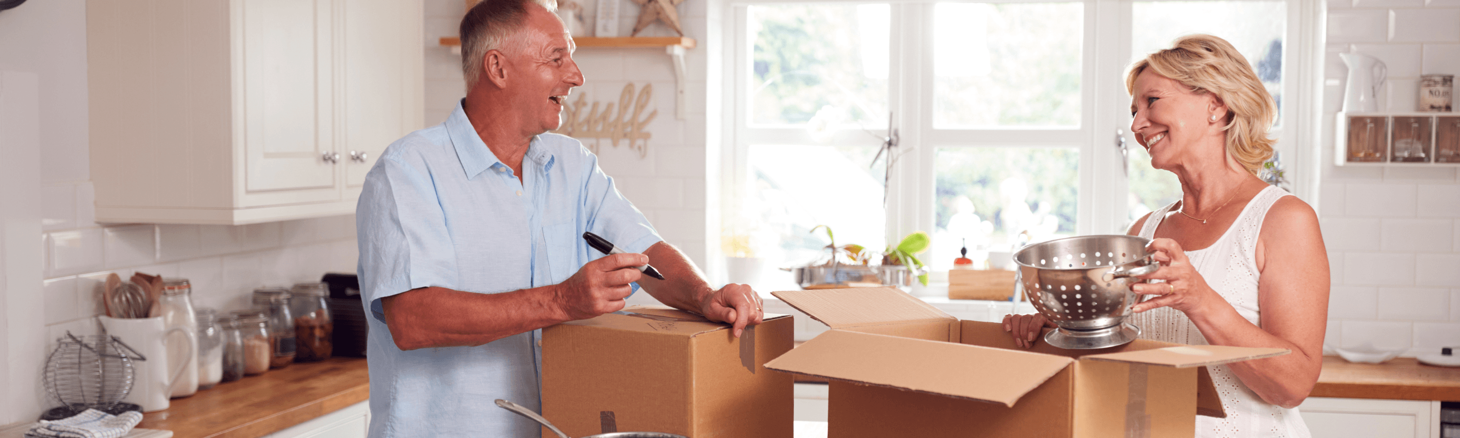 A couple is packing in a bright kitchen, one marking boxes while the other holds a colander and prepares to place it inside a box.
