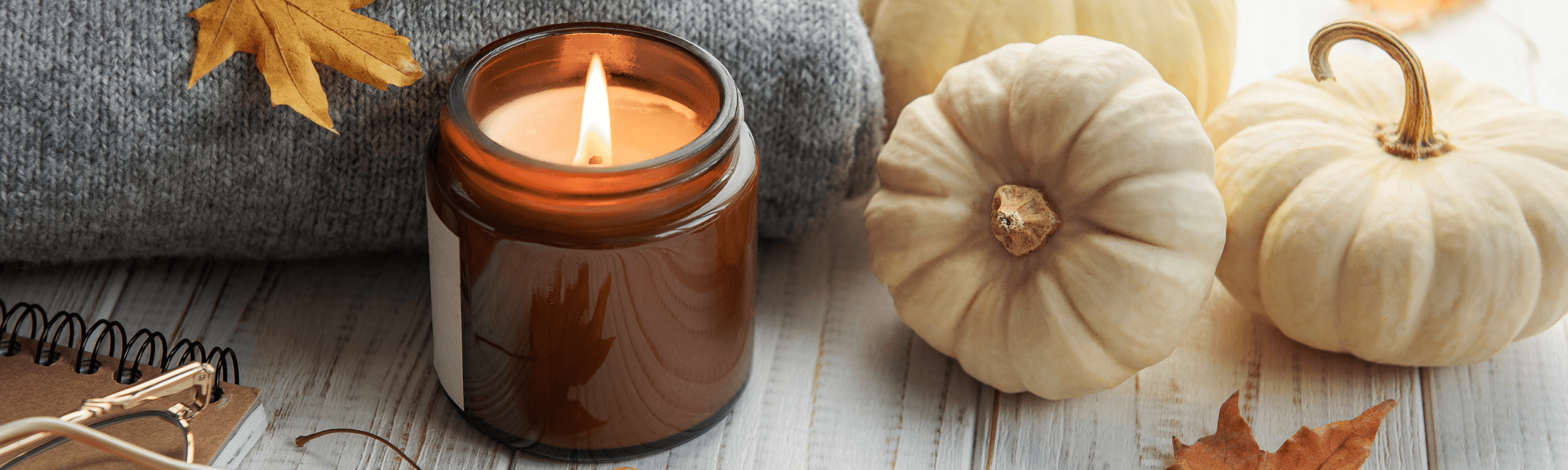 A lit amber jar candle sits next to white pumpkins, autumn leaves, and a cozy gray sweater on a wooden surface