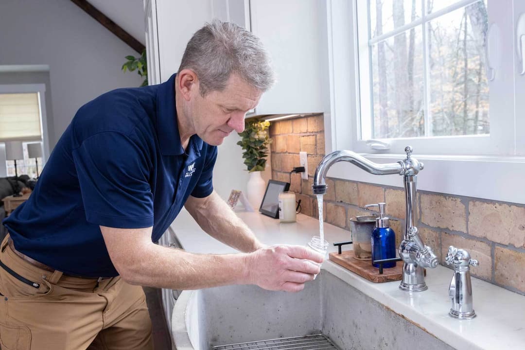 WIN Home Inspector filling a small container with water from a kitchen faucet in a bright, modern kitchen.