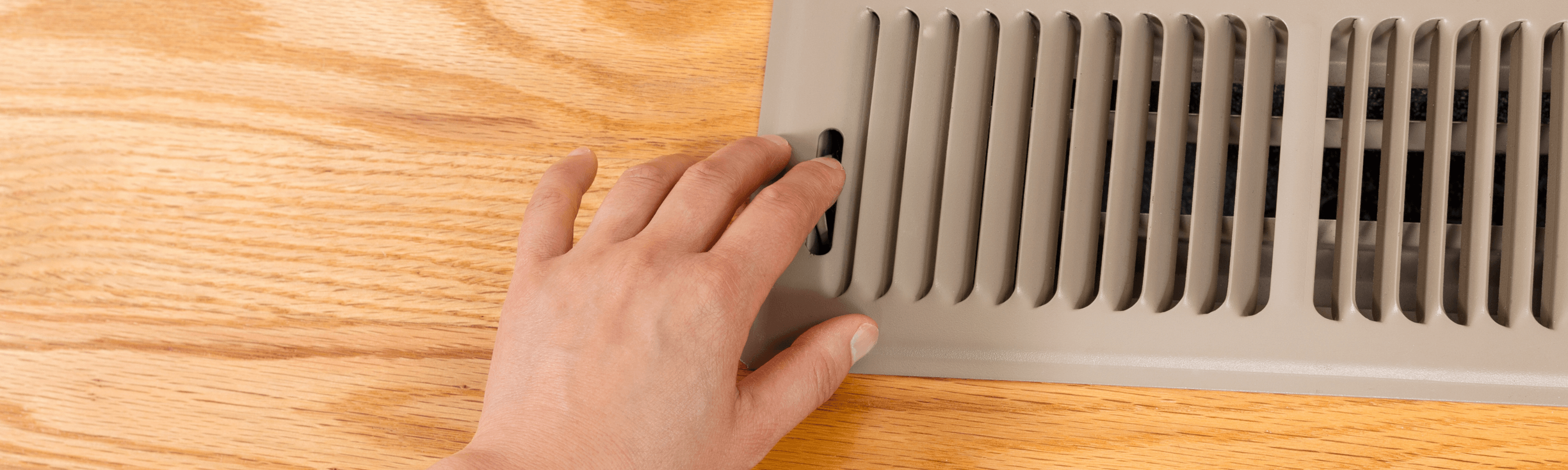 A hand adjusting a vent cover on a wooden surface, manipulating the slider to control airflow in a room.